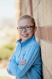 A photo of Morgan Yesnes leaning against a brick wall with his arms crossed and smiling. He is wearing a blue zip-up sweater and glasses. 